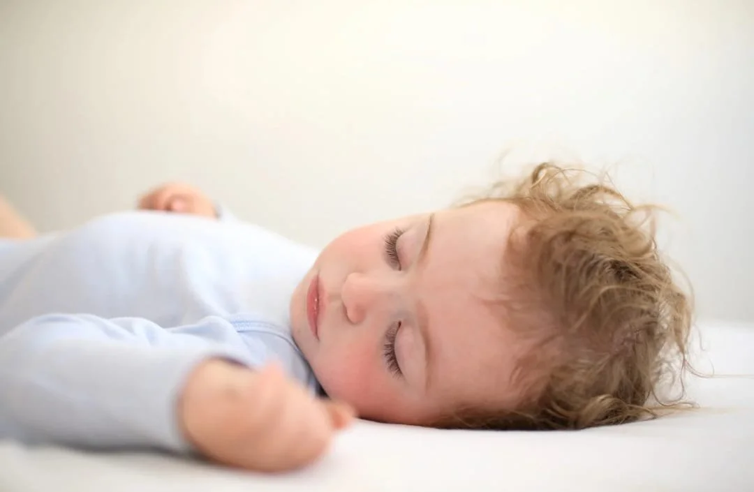 Bambino che dorme su un letto con un volto sereno, capelli ricci e occhi chiusi.