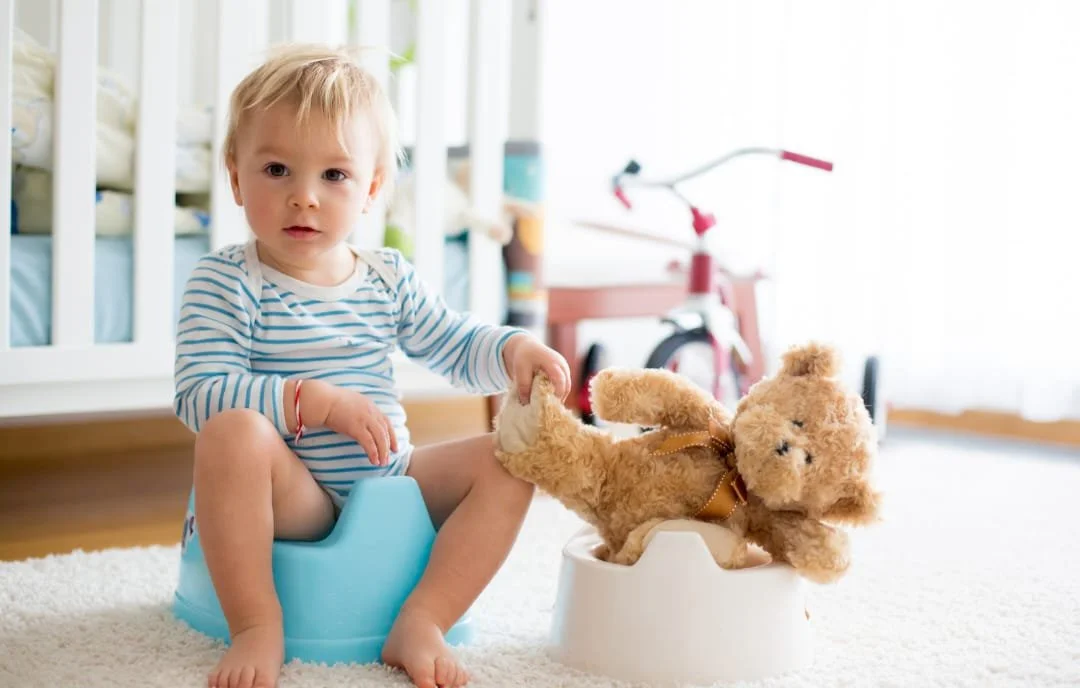 Bambino seduto su un vasino di plastica azzurro con un orsacchiotto di peluche sul bordo.
