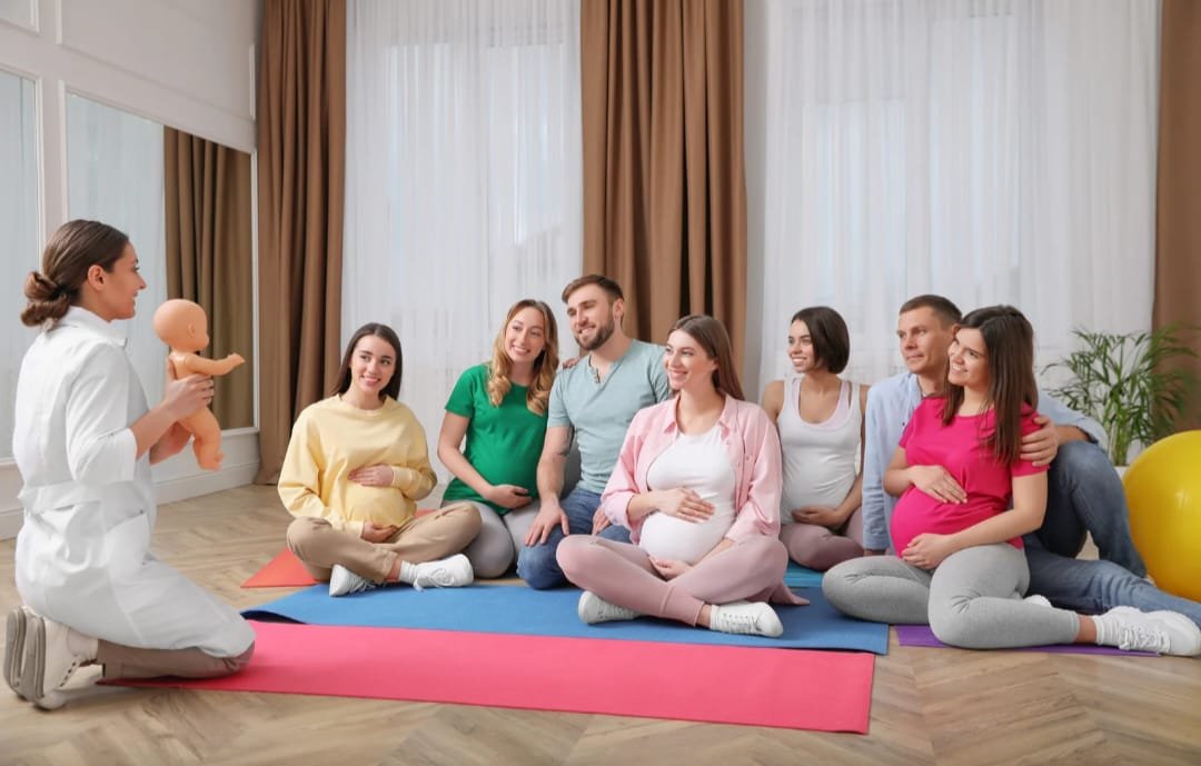 Gruppo di donne in gravidanza sedute su tappetini durante una lezione di preparazione al parto, con un'istruttrice che tiene un bambolotto in una stanza luminosa.