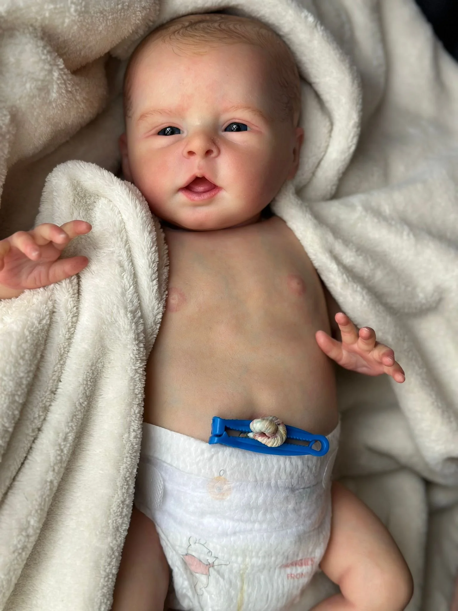 Neonato sdraiato su una coperta di peluche, con pelle chiara, occhi azzurri e bocca aperta, indossa un pannolino e una catenella colorata sulla pancia.