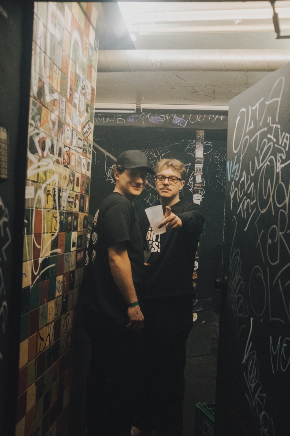 Raphi and Leon standing in a dark, graffiti-covered room, one smiling and the other pointing at the camera. One is wearing a cap and the other glasses, both dressed in black.