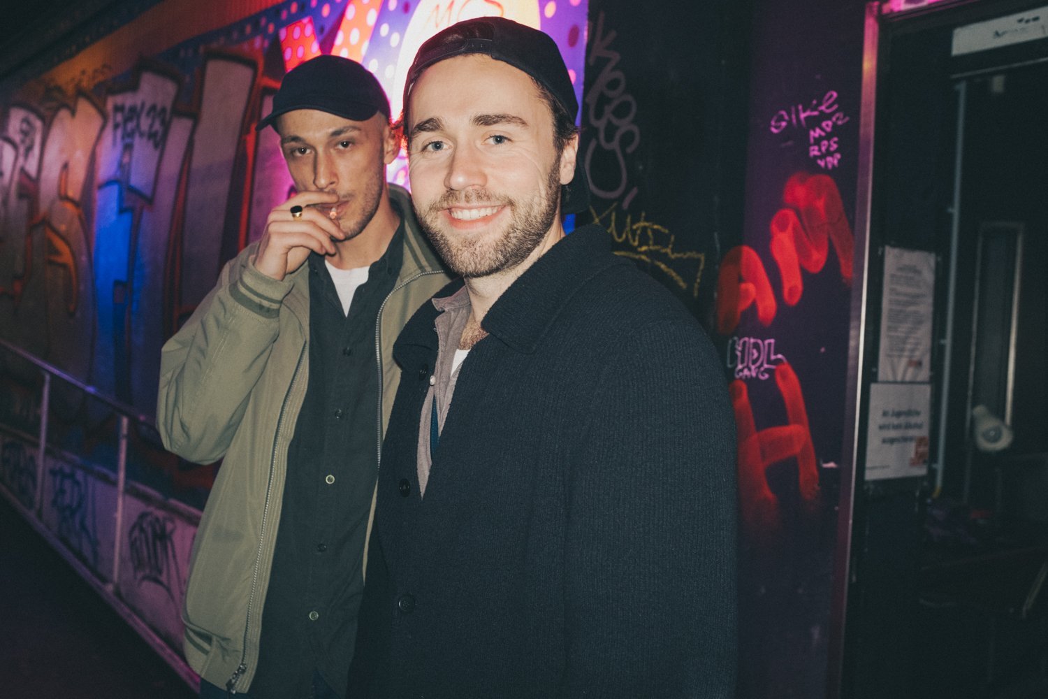 Fabi R. and Anel standing in a neon-lit, graffiti-covered lounge or club.