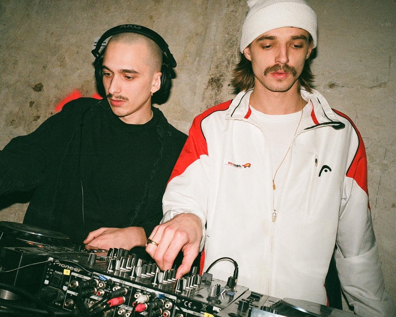 DJ. org and Ice Scholle with a mixer and headphones in front of a plain wall.