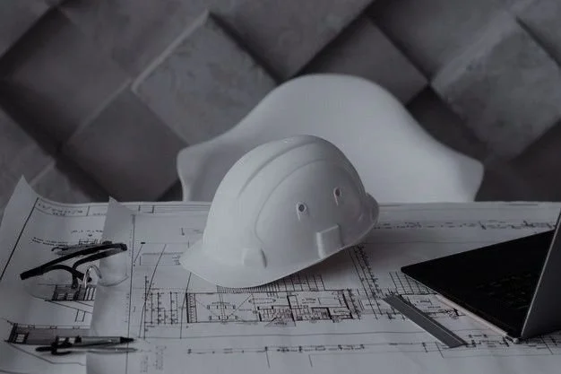 White construction helmet on architectural blueprints with tools and a laptop on a desk.