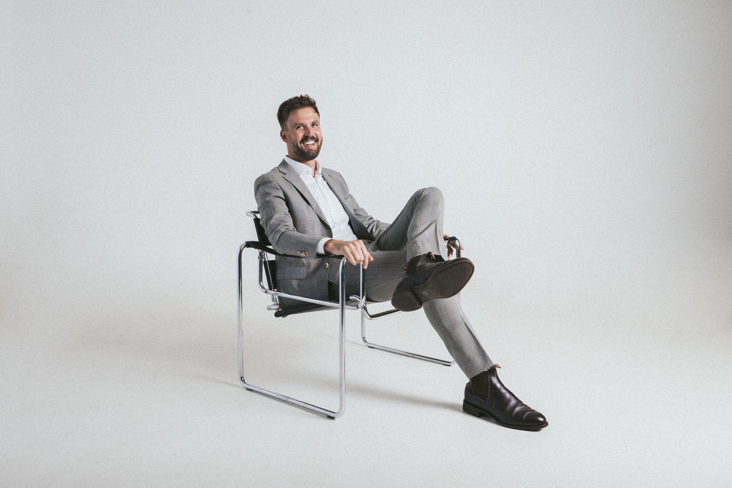 A man in a gray business suit sitting on a modern black chair, smiling, with one leg crossed over the other, against a plain white background.