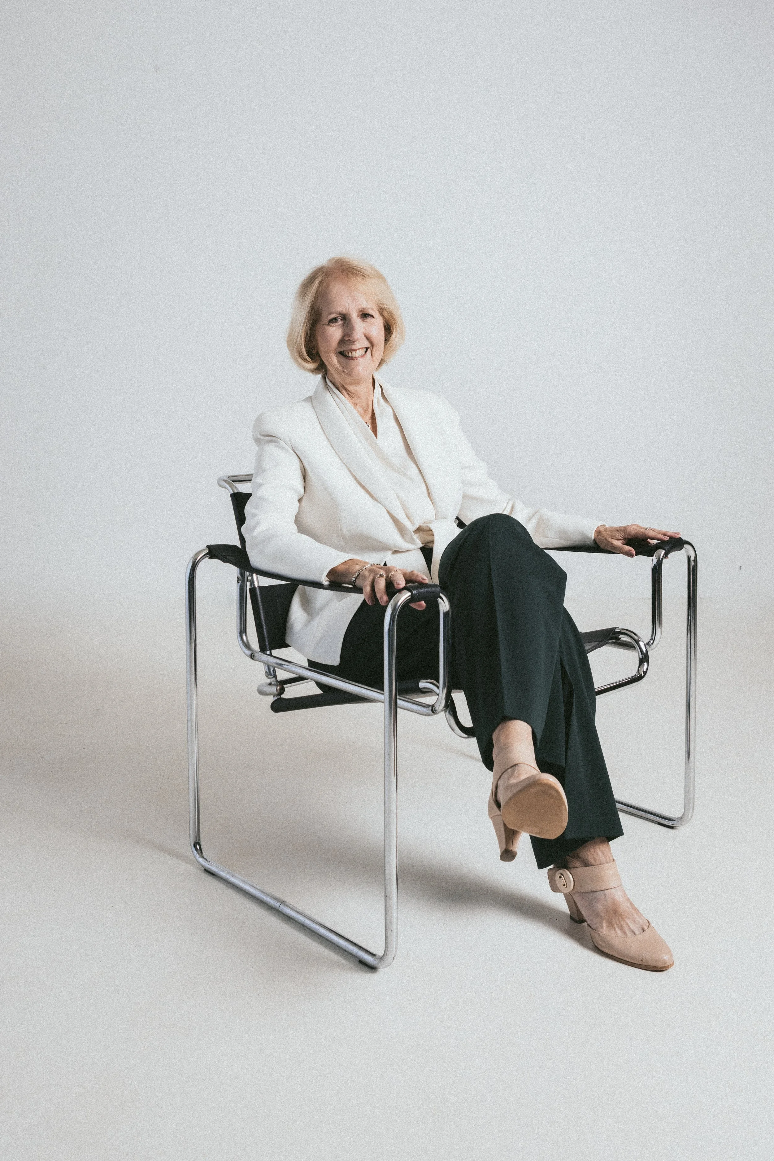 Smiling elderly woman in white blazer and black pants sitting on a modern black and chrome chair in a studio with plain white background.