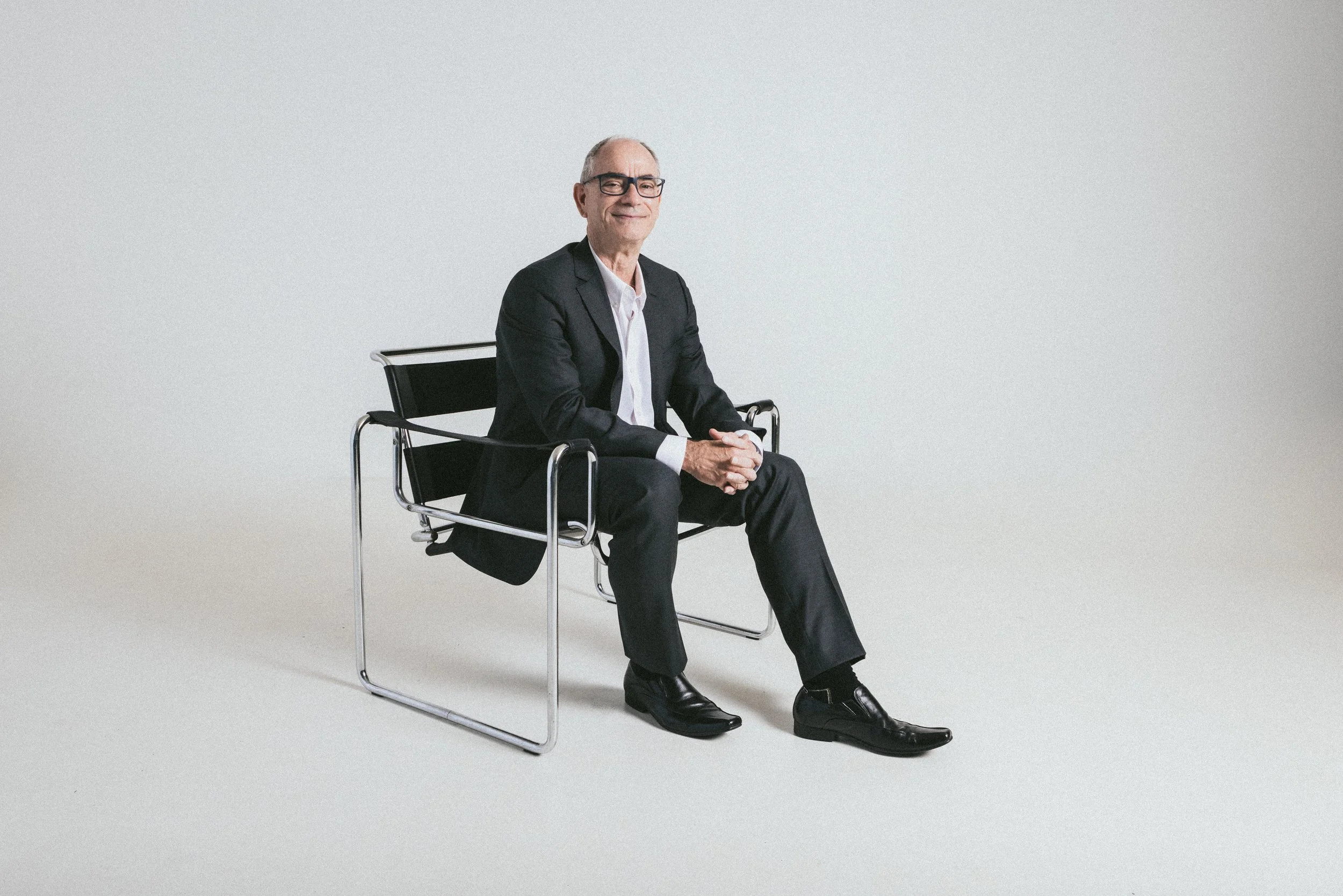 A man in a black suit and white shirt sitting on a modern black and chrome chair against a plain light background.