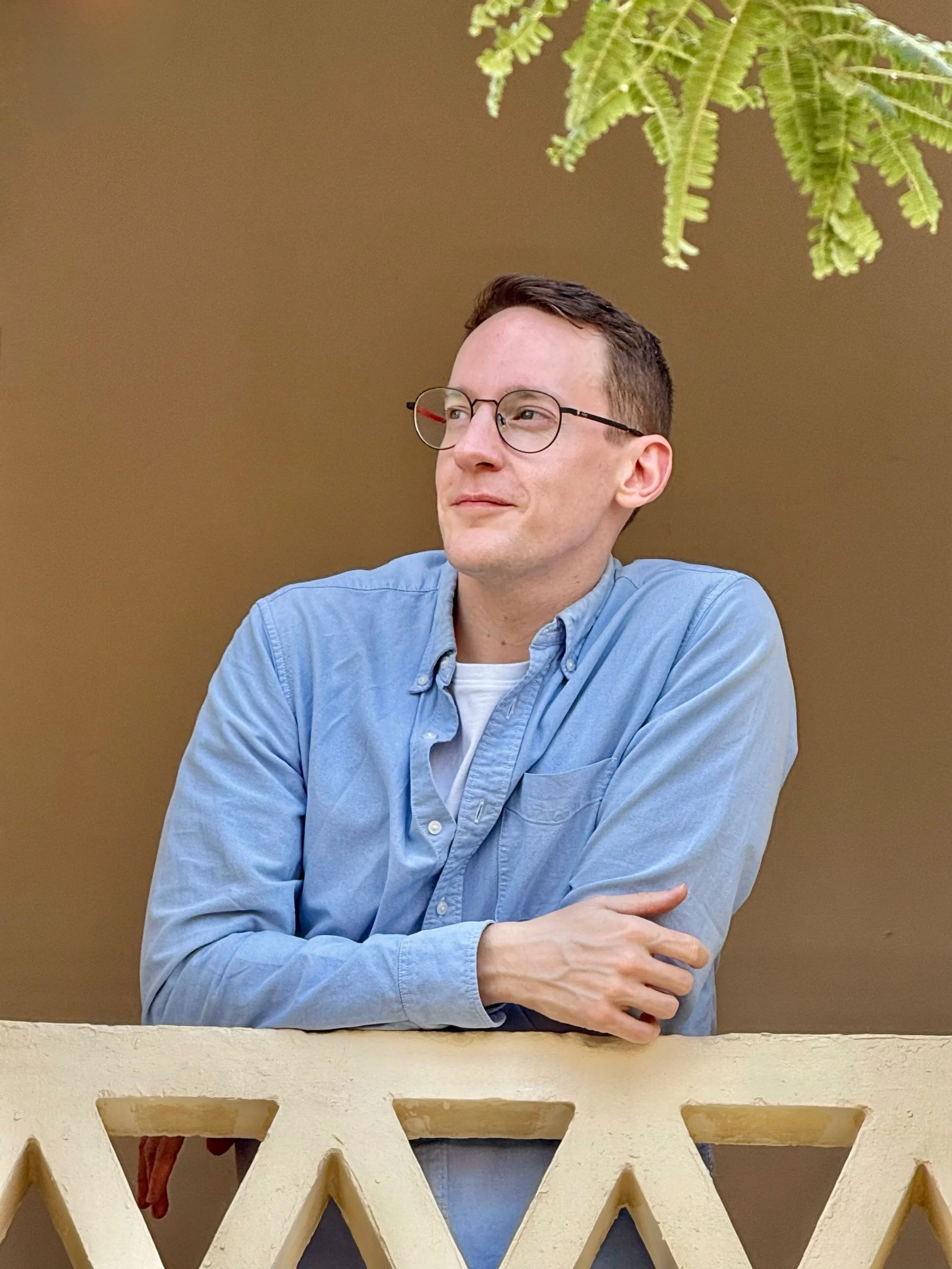 A man wearing glasses and a light blue shirt sitting on a balcony with decorative railing, looking to the side, with a green fern plant visible in the upper right corner.