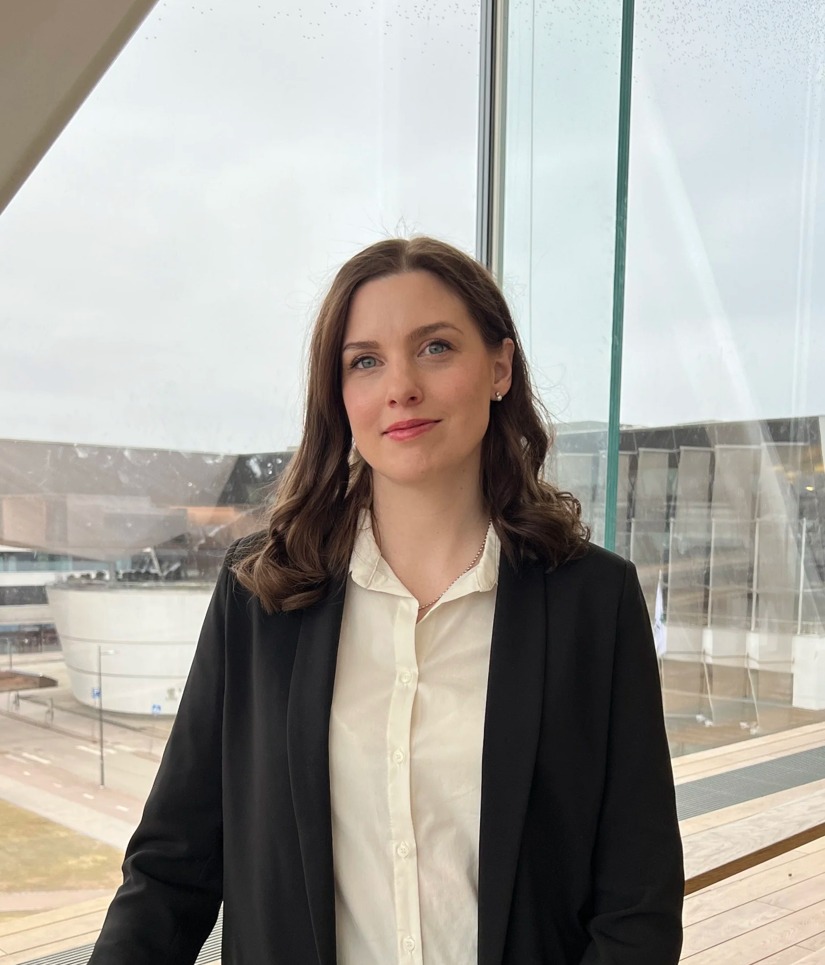 Kuva nainen, joka seisoo rakennuksen sisätiloissa, taustalla näkyy suuri ikkuna ja ulkona näkee ilmastonmuutoksen vaikutuksia, kuten pilvinen sää ja osittain jäinen pinta.