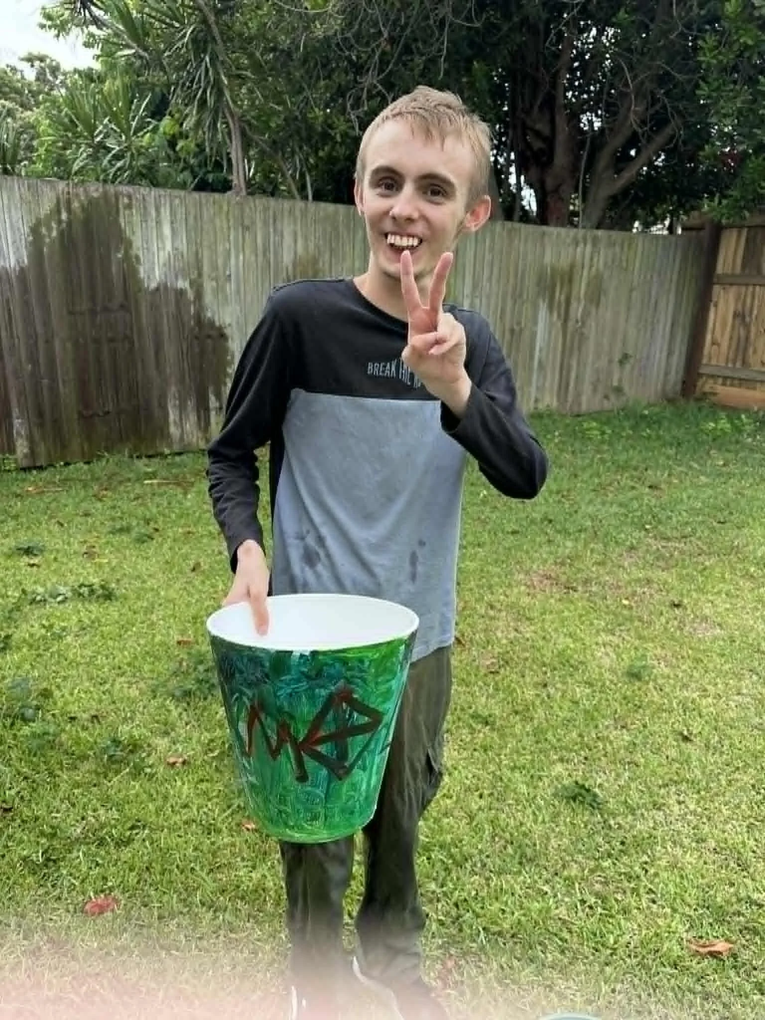 A young boy with short blonde hair, smiling and making a peace sign with his right hand, standing outside in a grassy backyard with a wooden fence and trees in the background, holding a colorful green bucket.