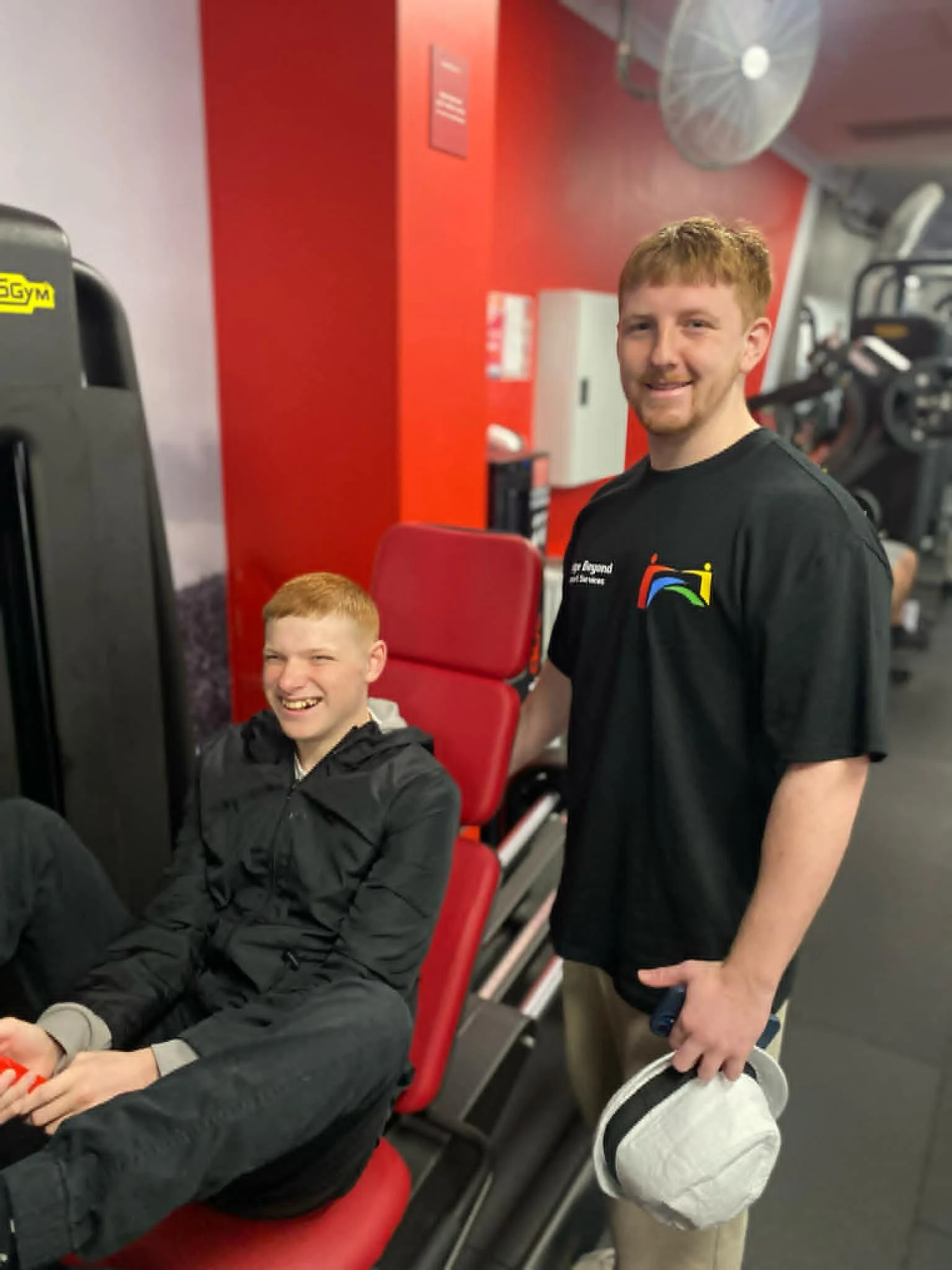 Two young men with red hair in a gym, one sitting on a leg extension machine and the other standing next to him. They are both smiling.