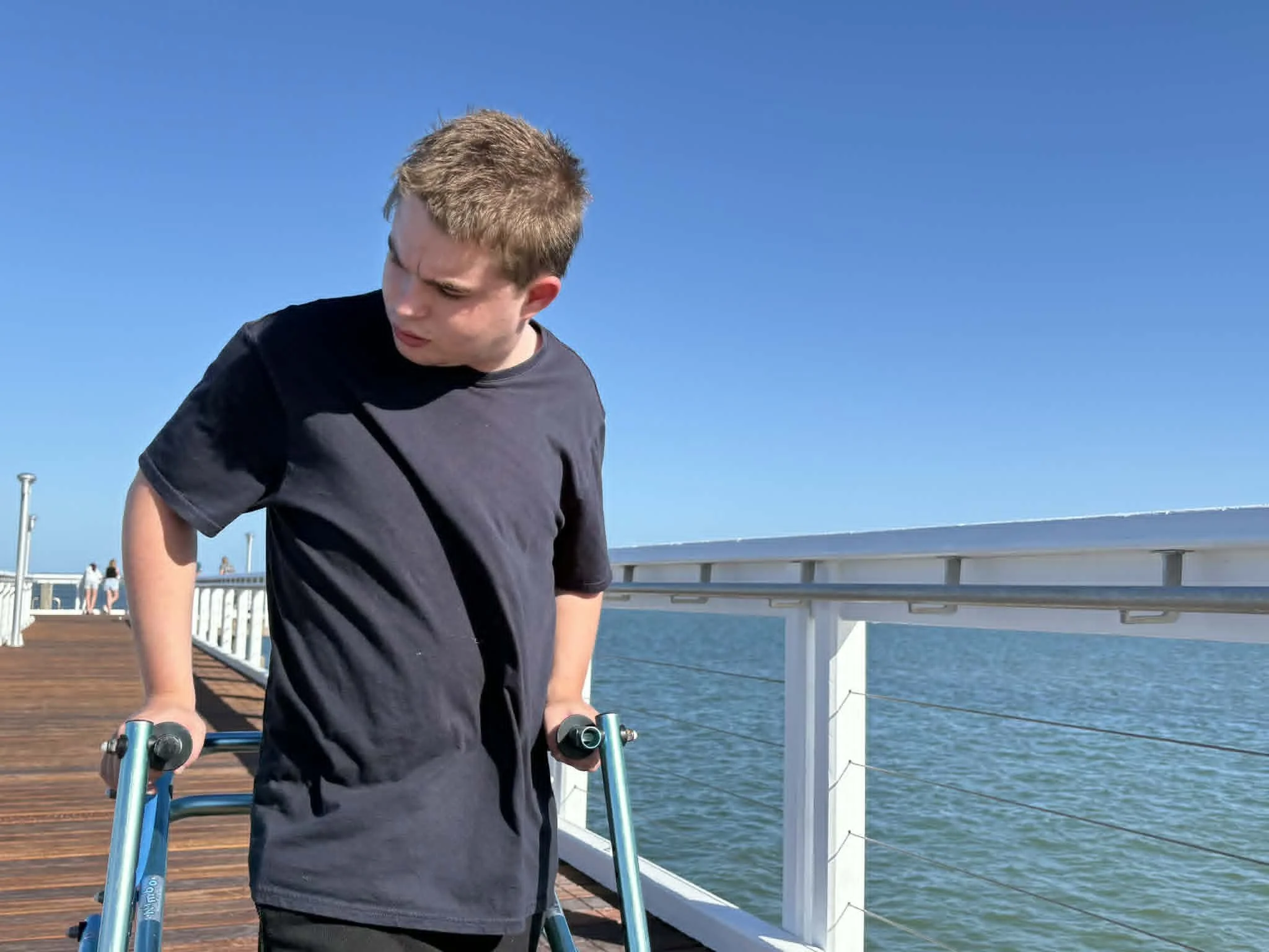 A young man with short, light brown hair using a walker on a wooden pier by the water under a clear blue sky.