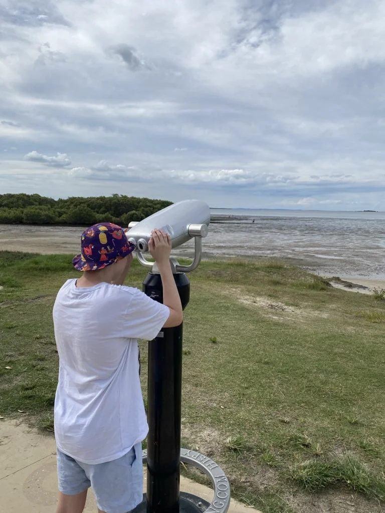 Man wearing a hat and white t-shirt looking through a coin-operated binocular viewer at a beach.