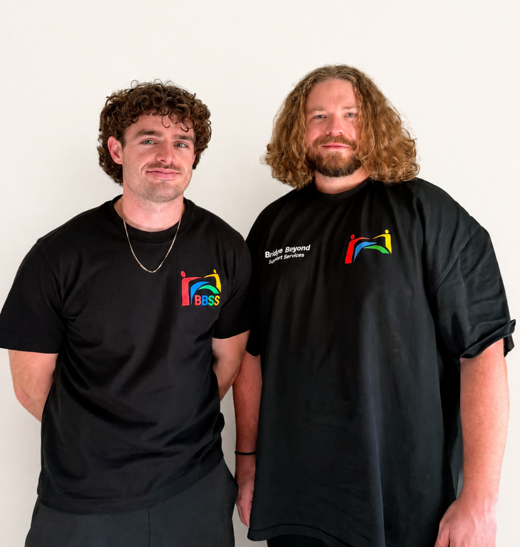 Two men standing side by side against a plain white wall, both wearing black t-shirts with colorful logos and text related to support services.
