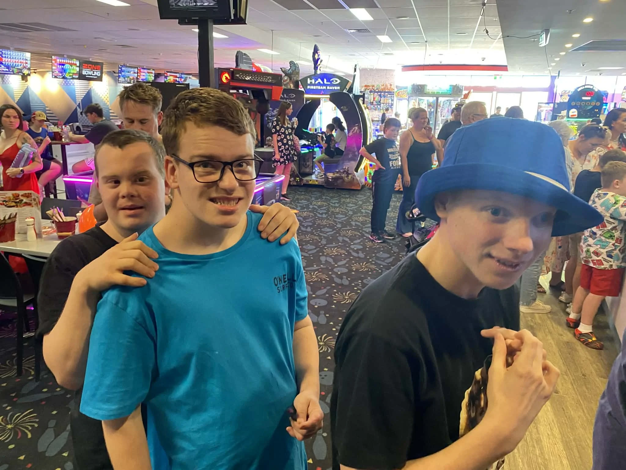 Two young men smiling and holding hands in an arcade, surrounded by people playing games and colorful arcade machines.