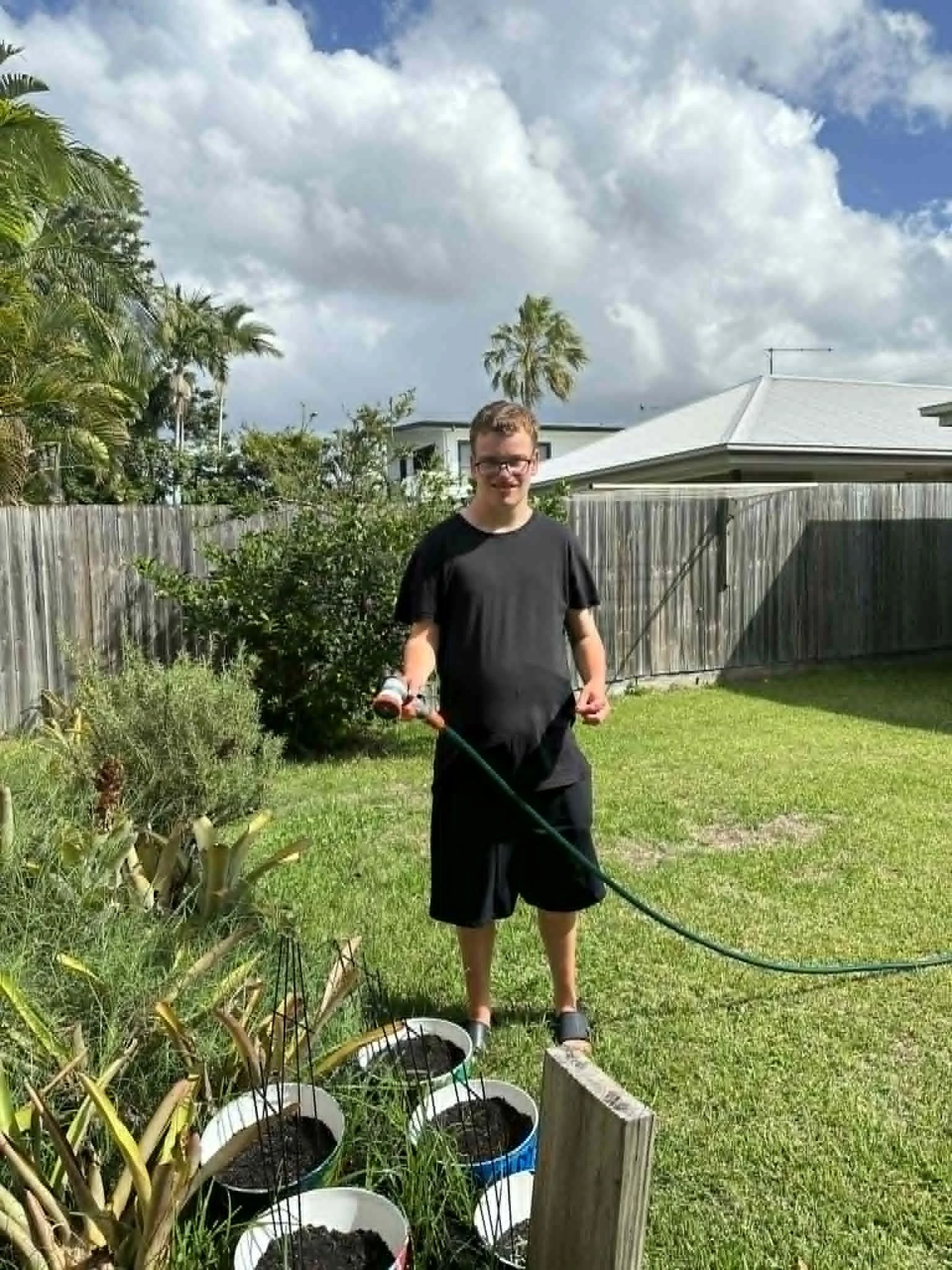 A young man with glasses, wearing a black T-shirt and shorts, standing in a backyard garden, holding a garden hose, with several pots of soil and plants around him, under a partly cloudy sky.