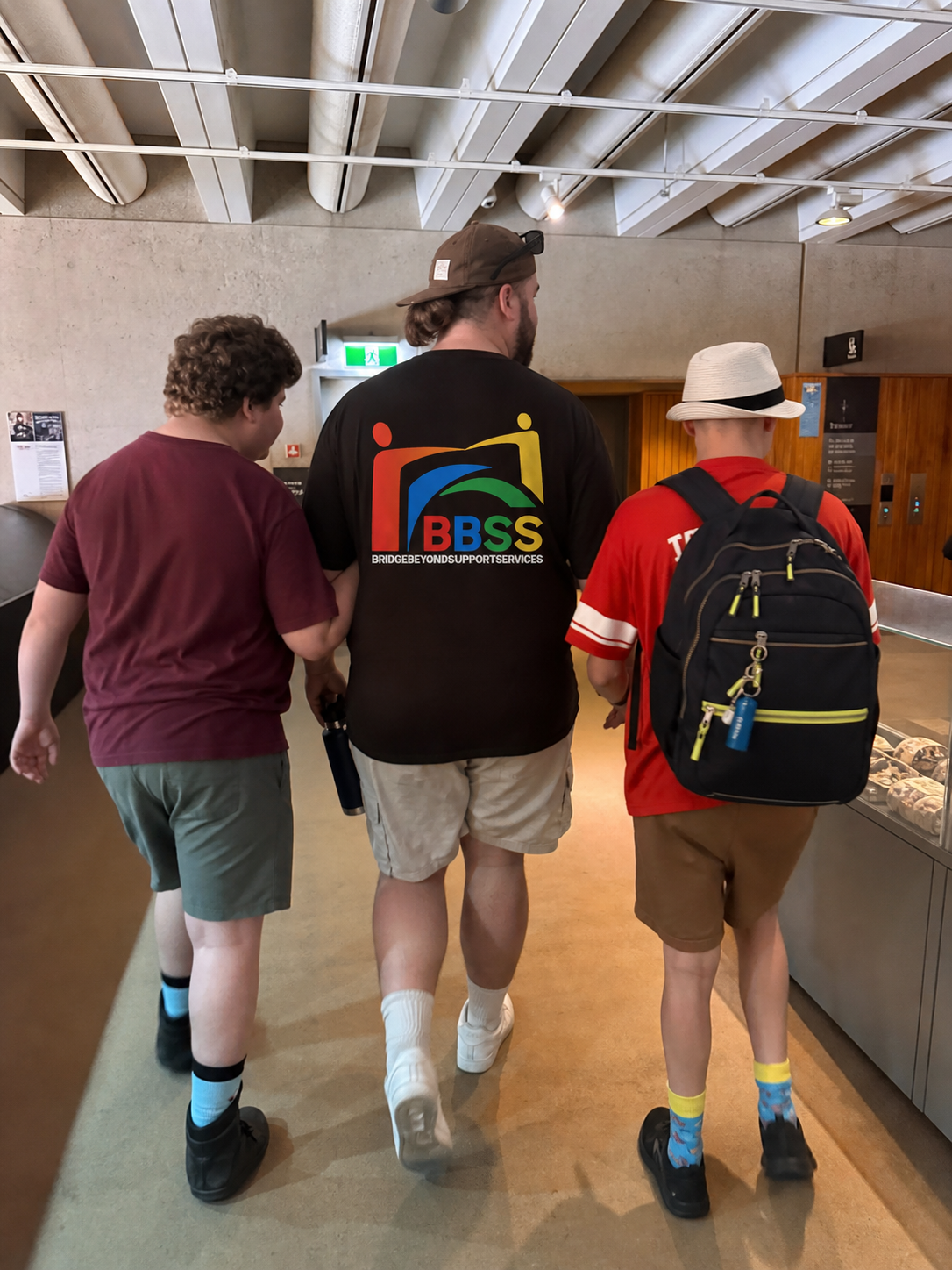 Three young men are walking in an indoor setting, with two on the sides and one in the middle. They are wearing casual clothing, shorts, and backpacks, and the middle one wears a black T-shirt with a colorful logo and the words 'BRIDGE BEYOND SUPPORT SERVICES.'