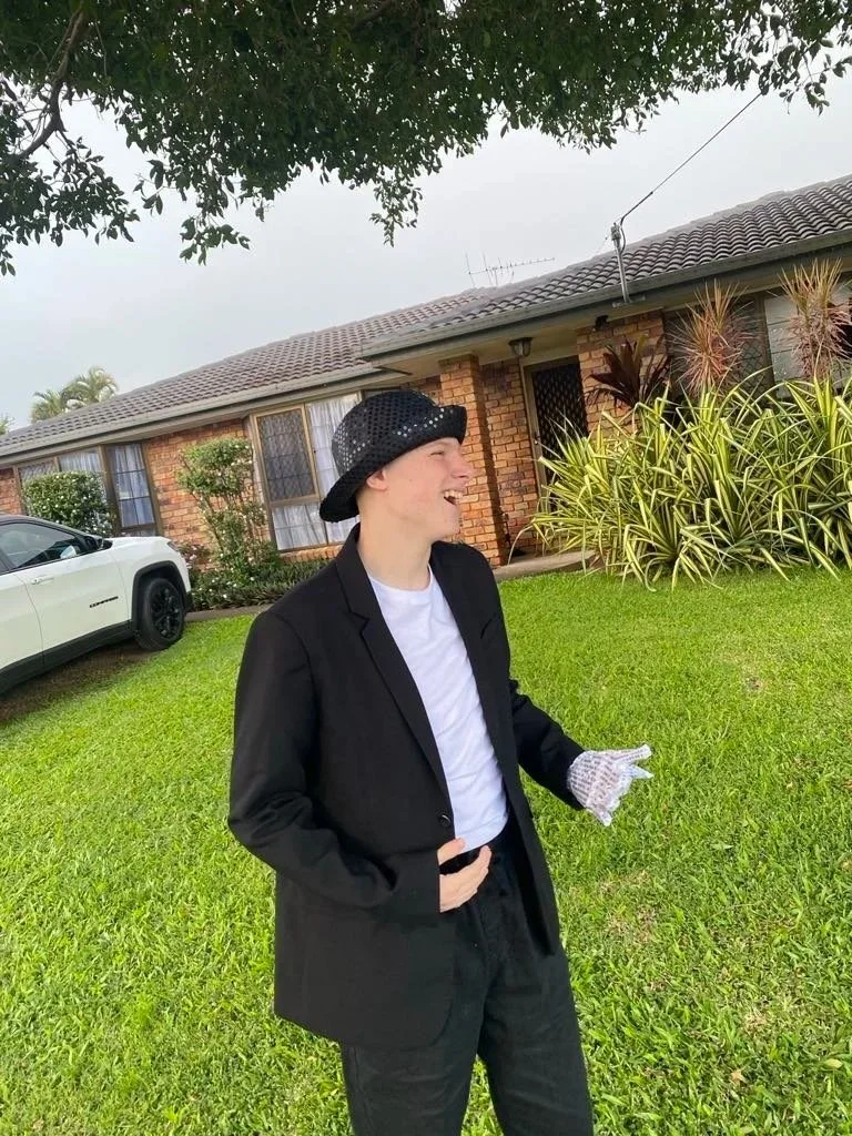A person imitating Michael Jackson wearing a black blazer, white t-shirt, polka dot hat, and a white, sparkly glove standing on a grassy lawn in front of a brick house with garden plants
