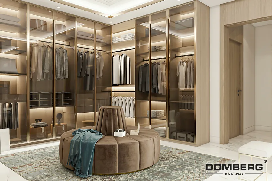 Luxurious walk-in closet with glass-front wardrobe cabinets filled with neatly hung clothes, folded linens, and accessories; central round seating with teal and brown fabric, surrounded by a patterned rug and beige tiled floor.