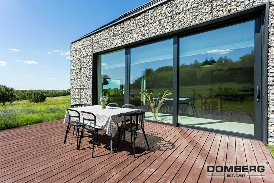 Outdoor patio with wooden decking, a dining table with a checkered tablecloth and four chairs, large sliding glass doors, and a modern building with textured stone exterior, overlooking a green landscape.