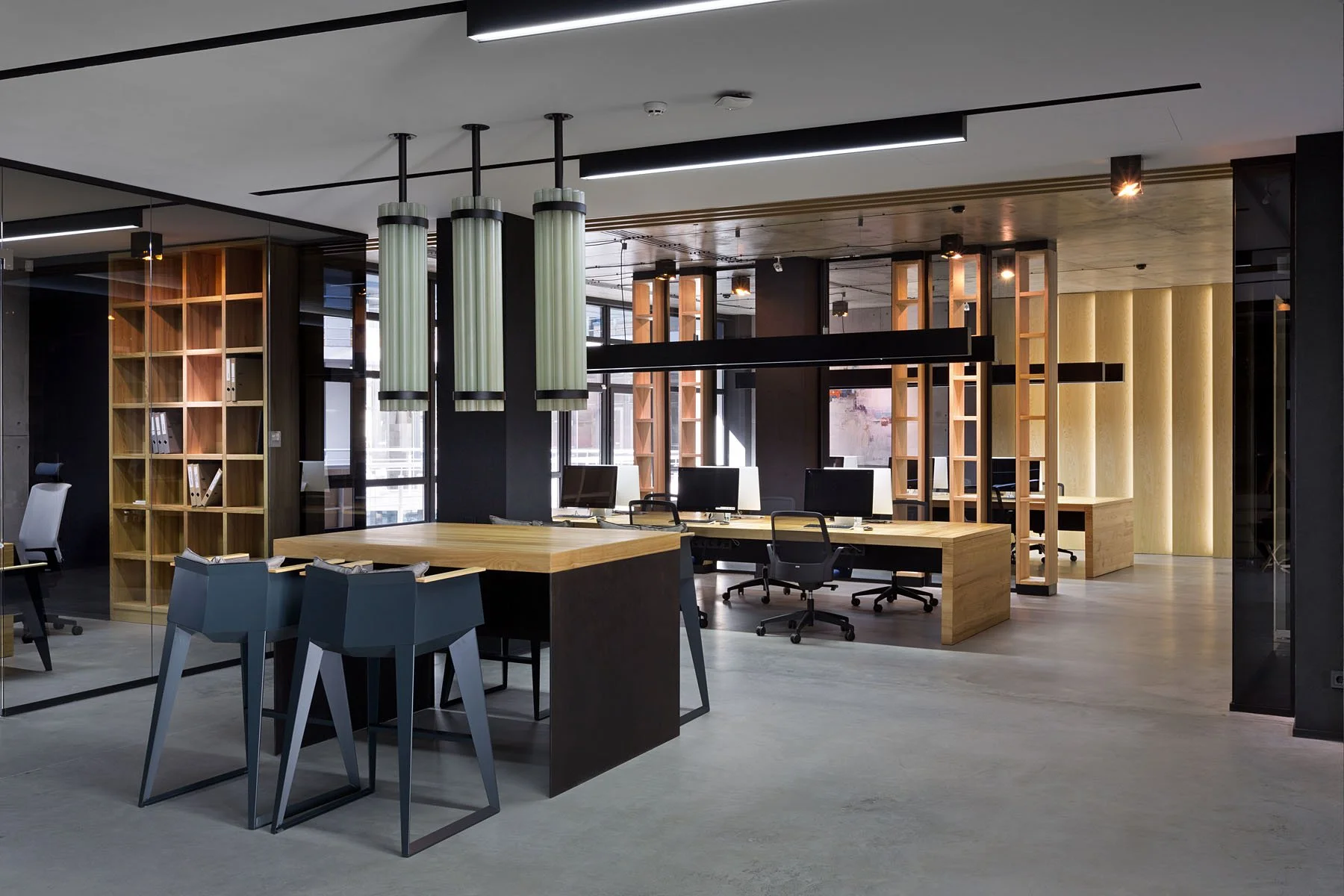 Modern office space with wooden desks, black chairs, computer monitors, wooden and black partitions, and hanging green light fixtures.