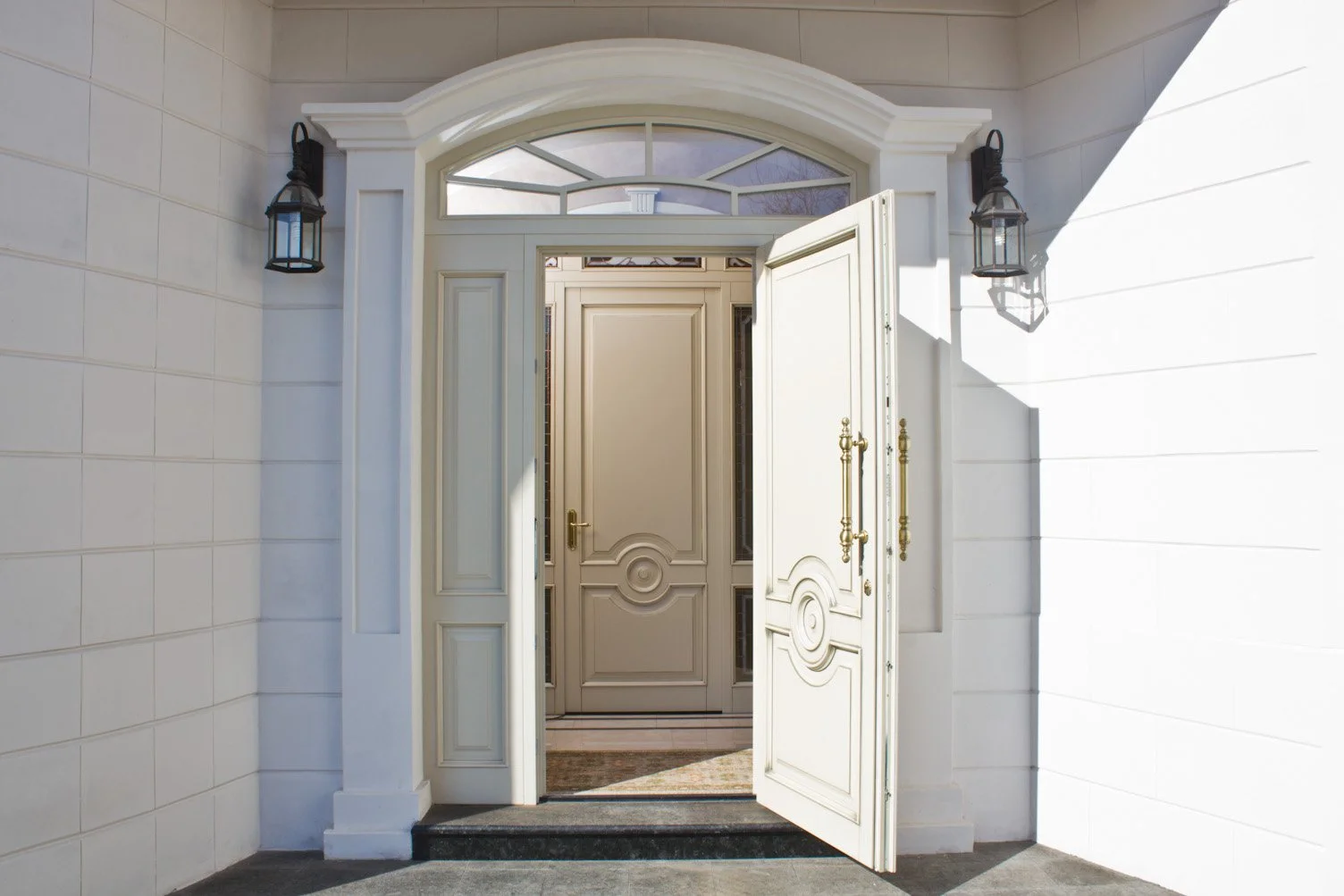 Open white front door with gold handles, flanked by two black lantern-style wall lights, on a white brick wall facade.