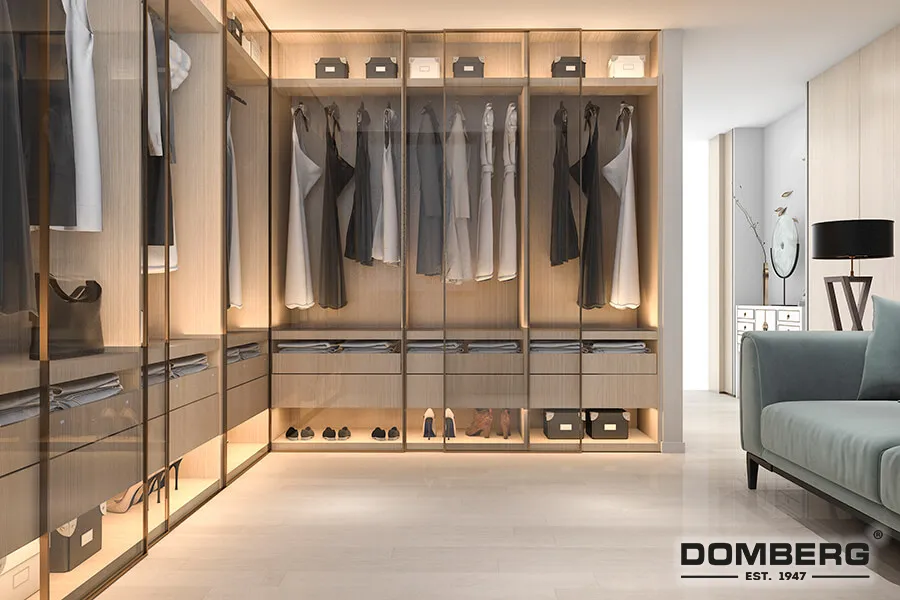 Modern walk-in closet with hanging clothes, shelves, and storage boxes, illuminated with soft lighting, adjacent to a light-colored sofa and side table.