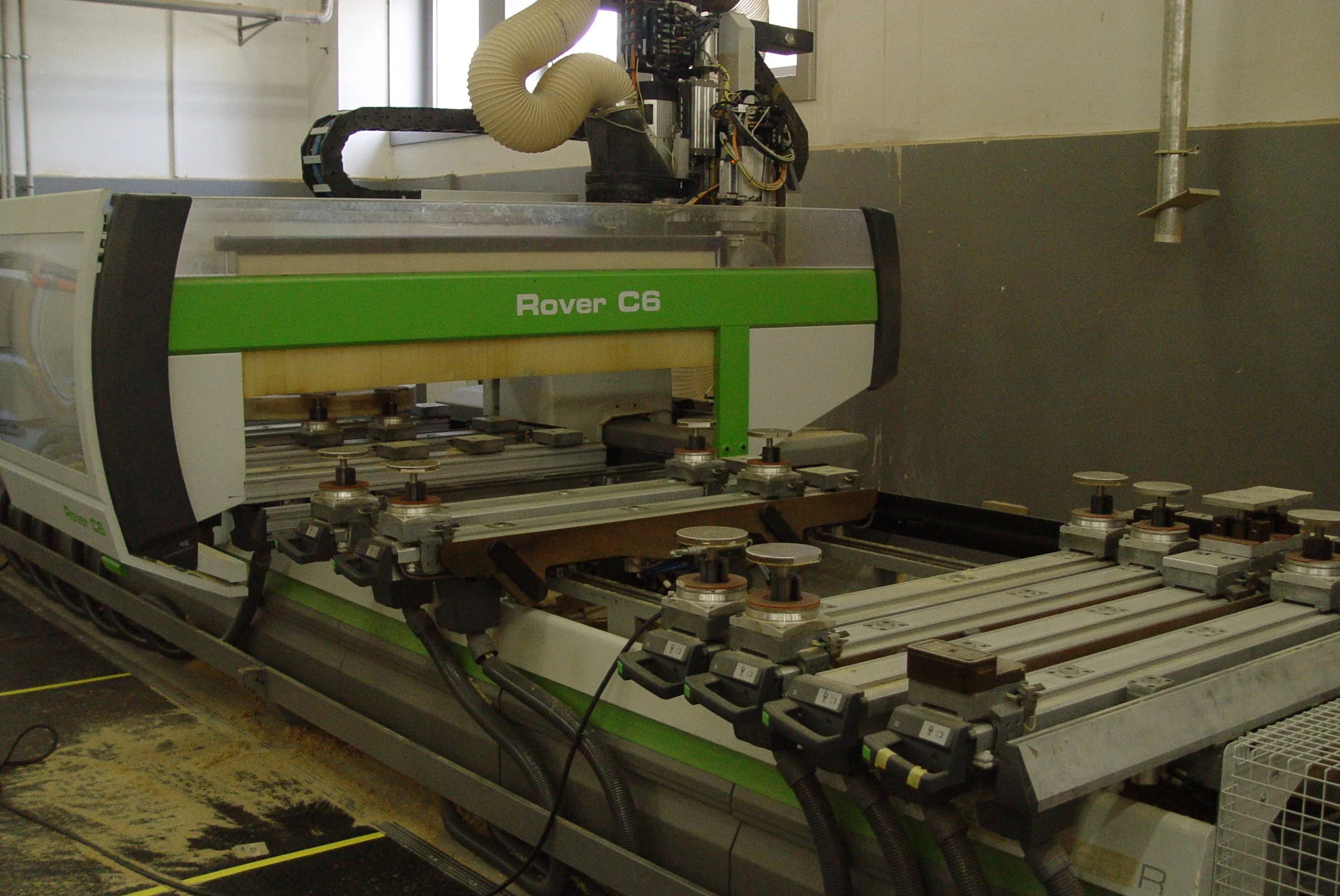 A large industrial machine labeled 'Rover C6' in a workshop or manufacturing environment. The machine has multiple components, including vacuum hoses and metal parts, with a green and white color scheme. The surrounding area includes a floor with marked safety lines and some equipment.