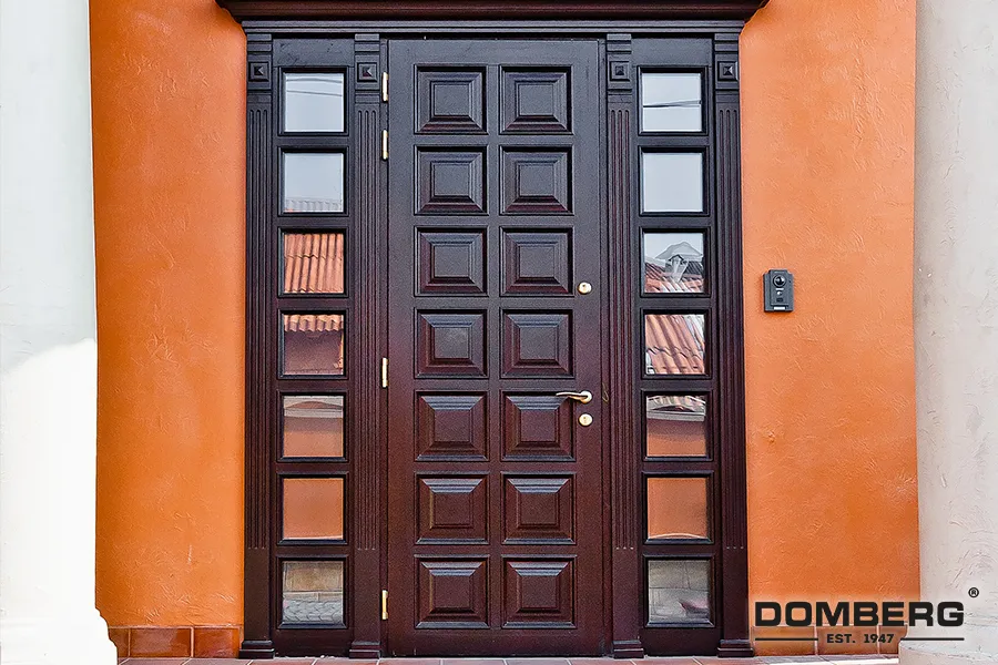 Dark wooden front door with geometric panel design, flanked by glass panels, and an orange stucco wall with a doorbell on the right.