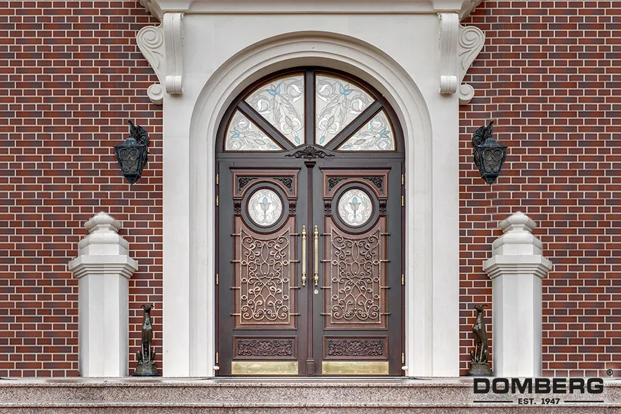 A pair of ornate wooden double doors with decorative wrought iron grills and brass handles, set within a white stone archway, flanked by brick walls and decorative statues on steps.