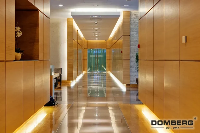 Modern hotel or office building hallway with wooden panel walls, polished wooden floor, and ambient lighting along the baseboards, leading to glass doors at the end.