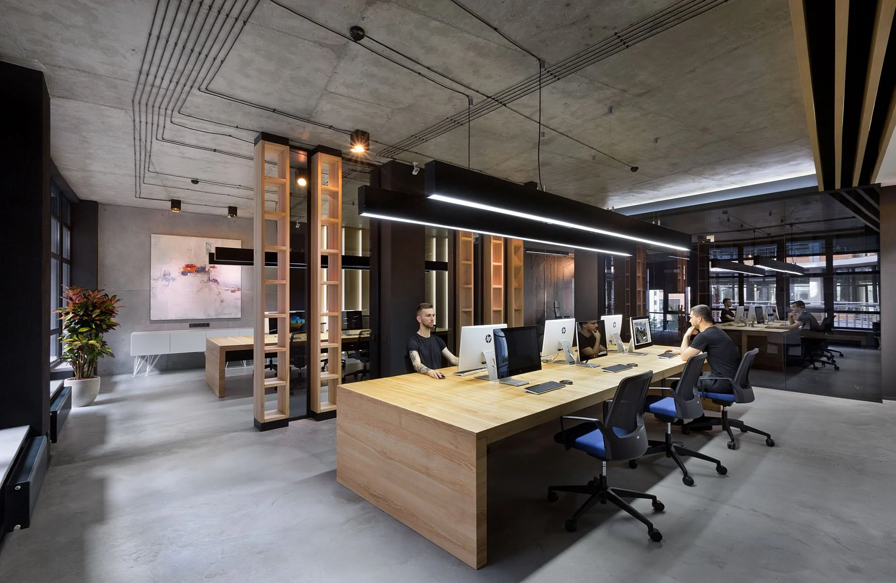 Modern office space with several employees working at desktop computers at a large wooden table, with black and wood paneling, large windows, and industrial-style lighting.