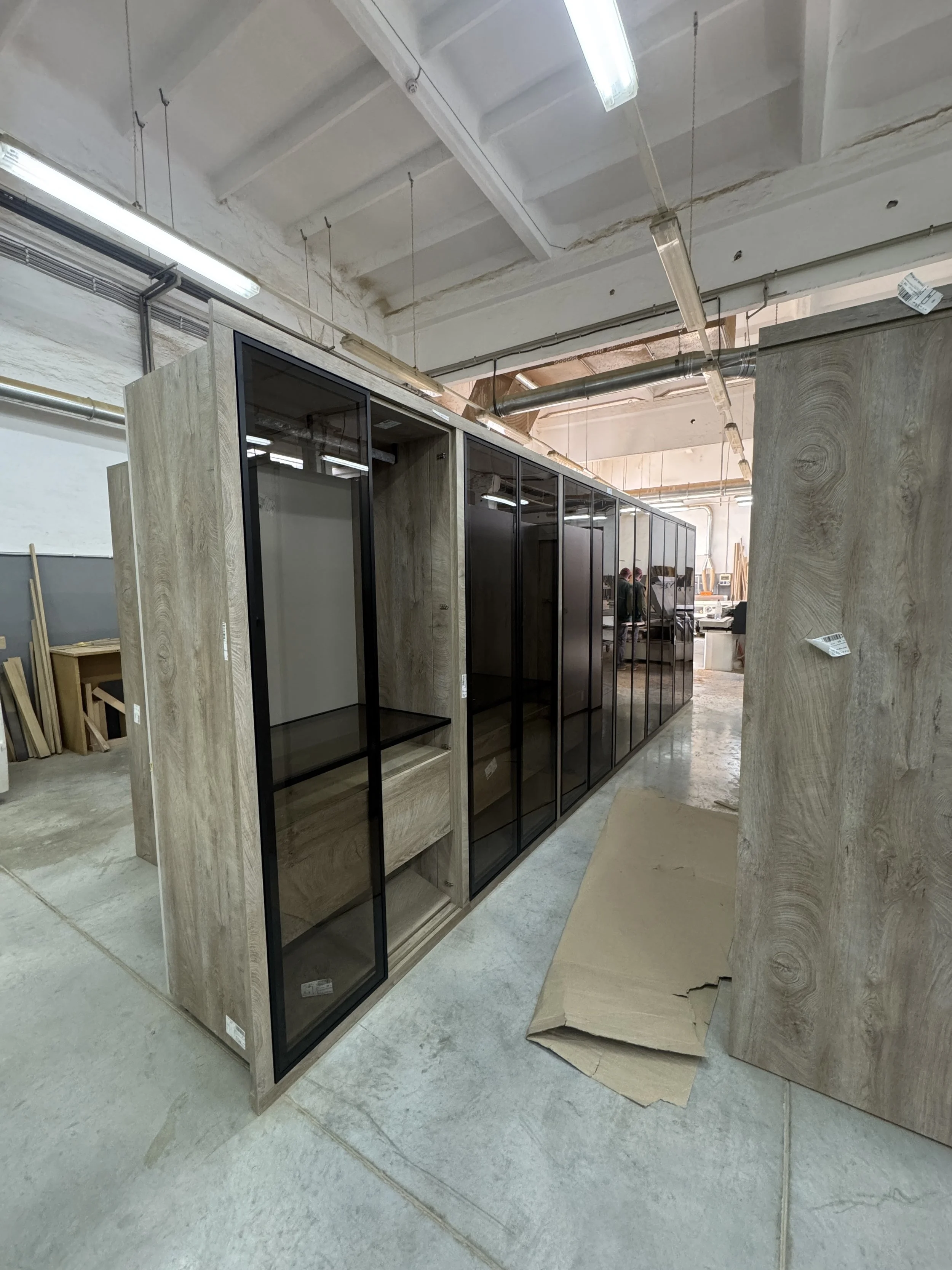 Storage fixtures in a warehouse or showroom with wooden and glass display cabinets, two workers in the background, and unfinished ceiling with fluorescent lights.