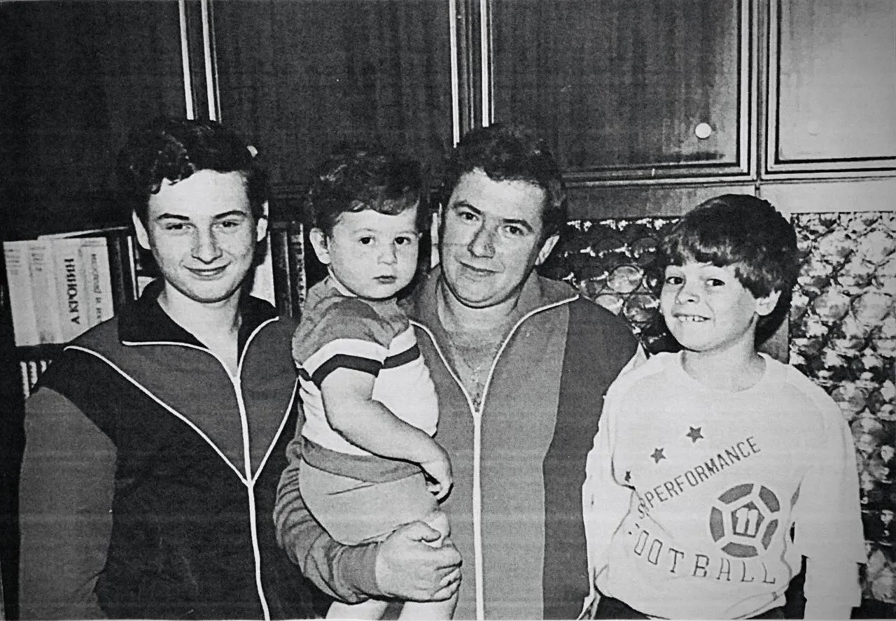Black and white photo of four males, a young man on the far left, an older man holding a small child in the center, and a boy on the far right, standing in front of bookshelves and wallpaper.