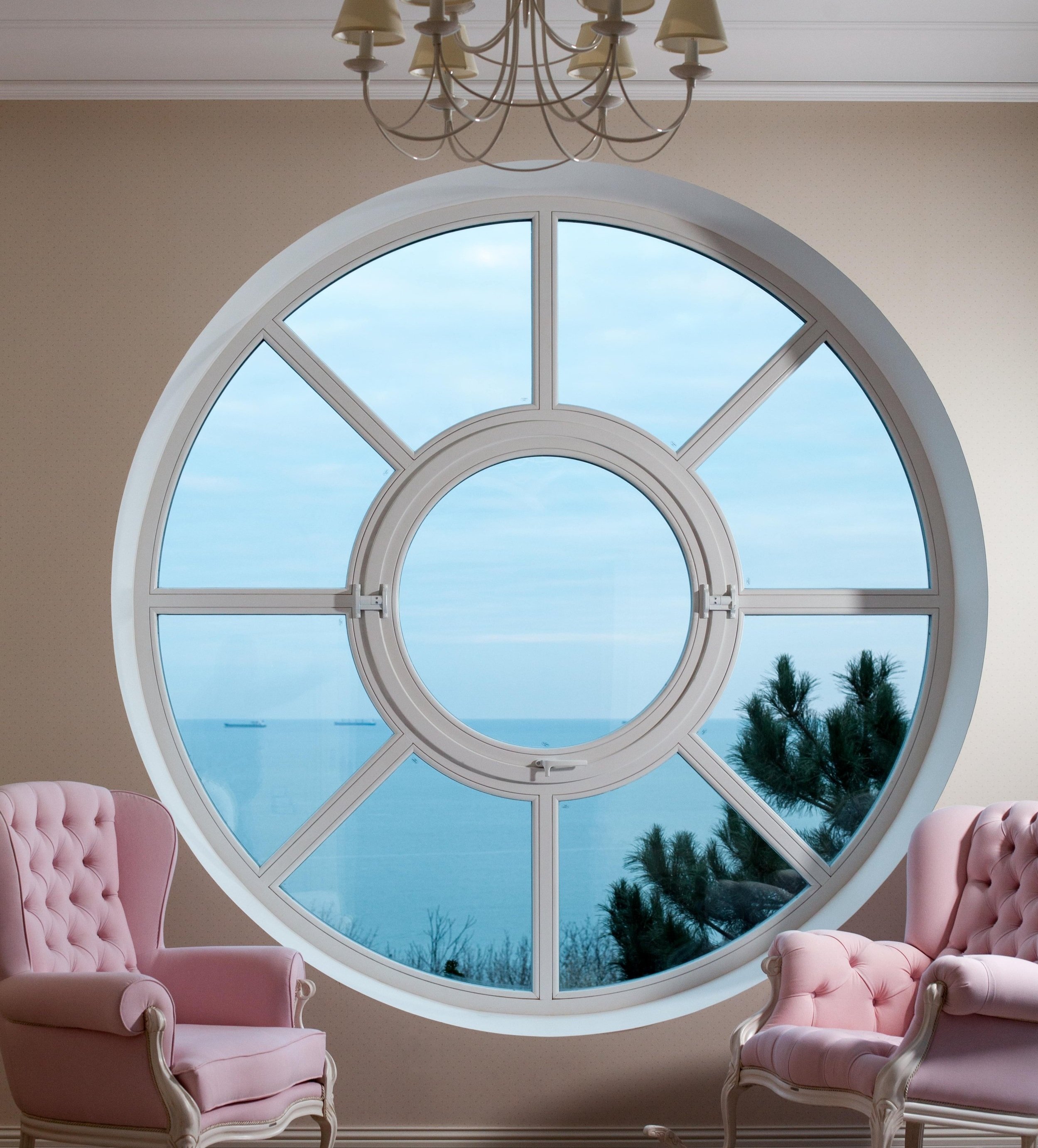 A large circular window with a view of the ocean and a cloudy sky, surrounded by two pink upholstered chairs, and a chandelier hanging from the ceiling.