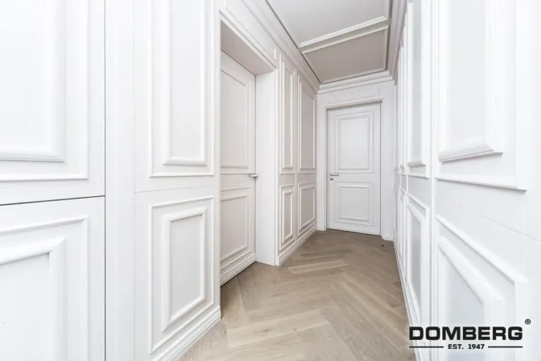 A narrow hallway with white paneled walls and crown molding, wooden flooring, and closed white doors.