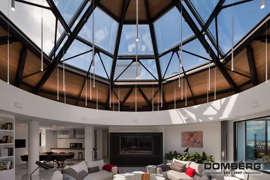 Interior of a modern living room with a large circular skylight ceiling with glass panels and a view of the sky. The room features a white sofa with cushions, a fireplace, and a dining area in the background. There are plants, artwork, and large windows allowing natural light in.