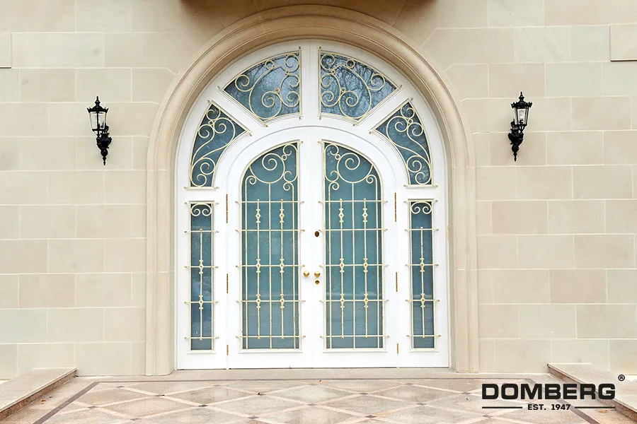 Front entrance with a large white arched iron gate with decorative scrollwork, flanked by stone walls and black lantern-style wall sconces.