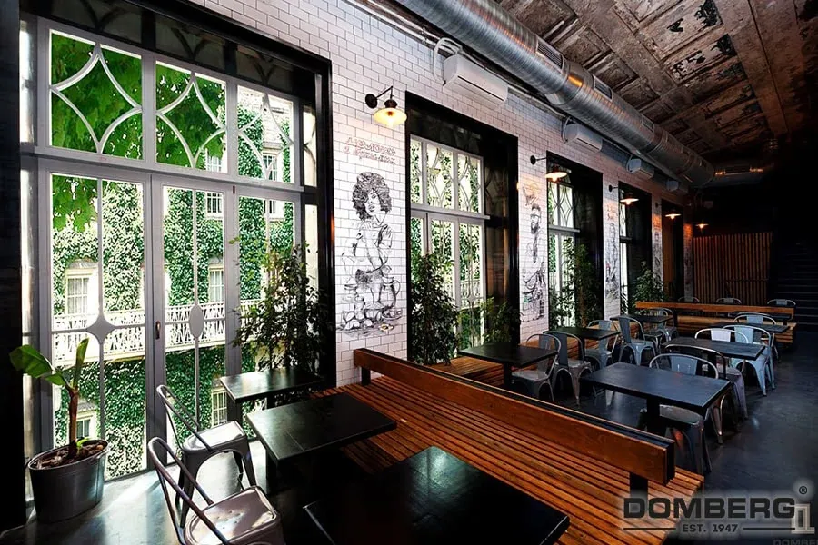 Interior of a modern restaurant or café with large windows, white brick walls decorated with black and white graffiti art, potted plants, and black tables with metal chairs.