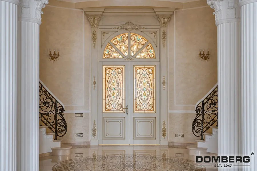Elegant entrance with a double door featuring ornate stained glass, surrounding decorative trim, flanked by two classical columns, with a marble floor and wall sconces.