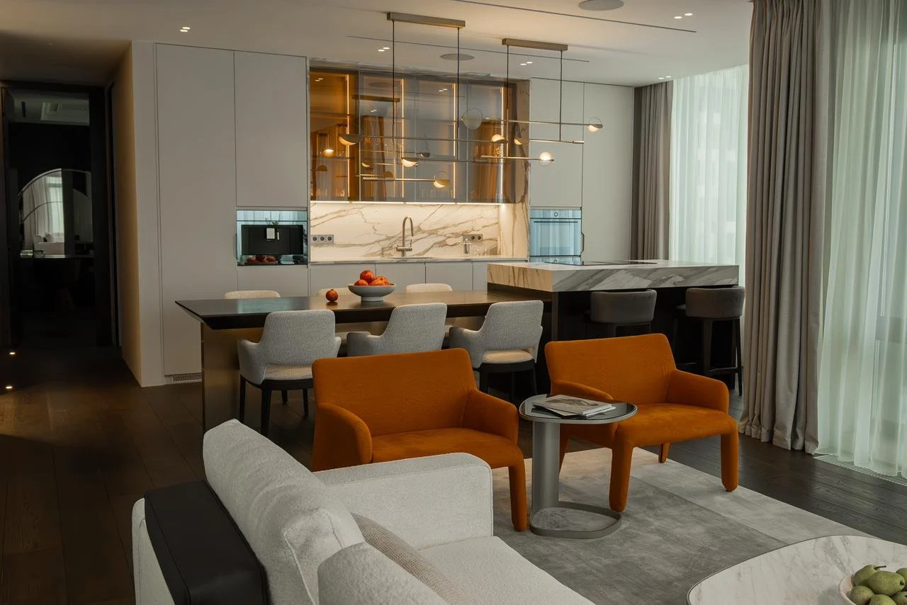 Modern living room with orange armchairs, white sofa, small side table with magazines, and a dining area with white chairs and a dark table in an open-concept space. Elegant kitchen in the background with marble countertops, hanging light fixtures, and large windows with curtains.