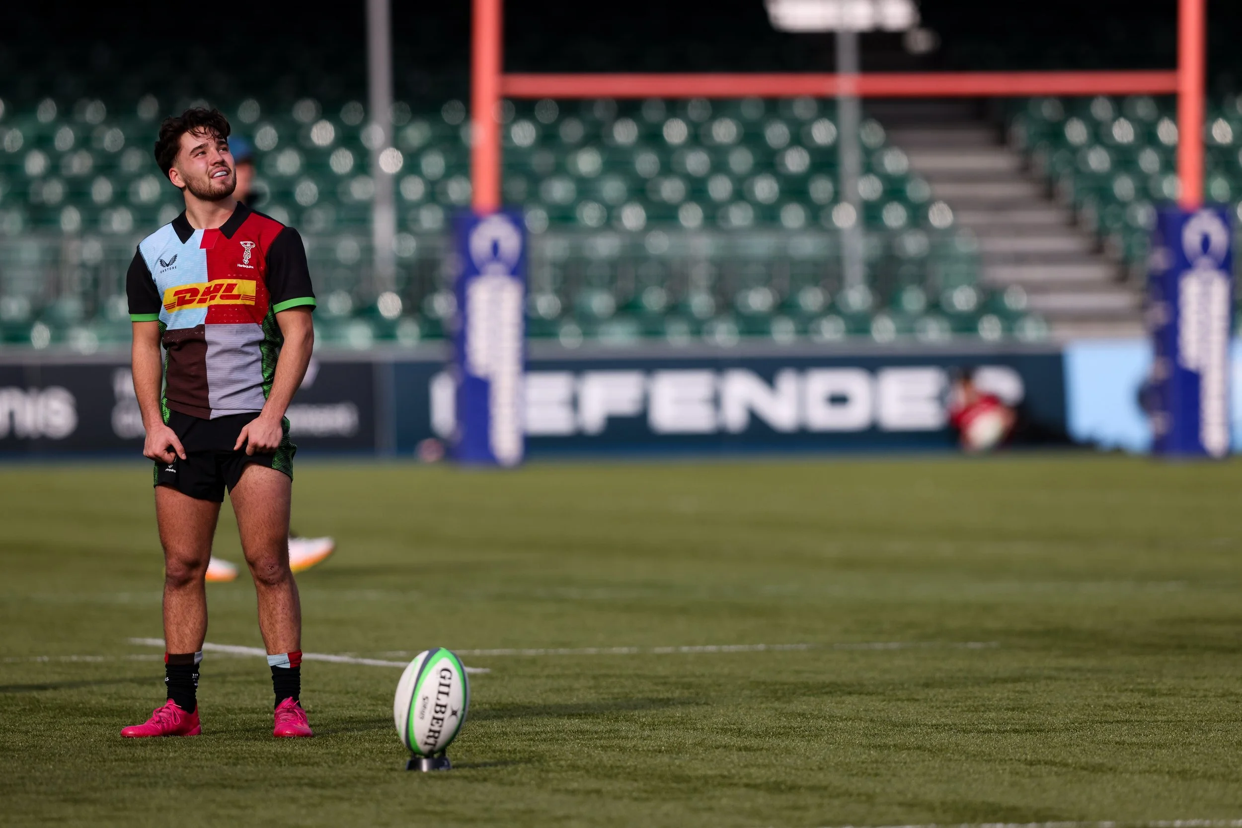 A rugby player standing on a field with a rugby ball nearby, wearing a colorful jersey and black shorts, with empty stadium seats in the background.