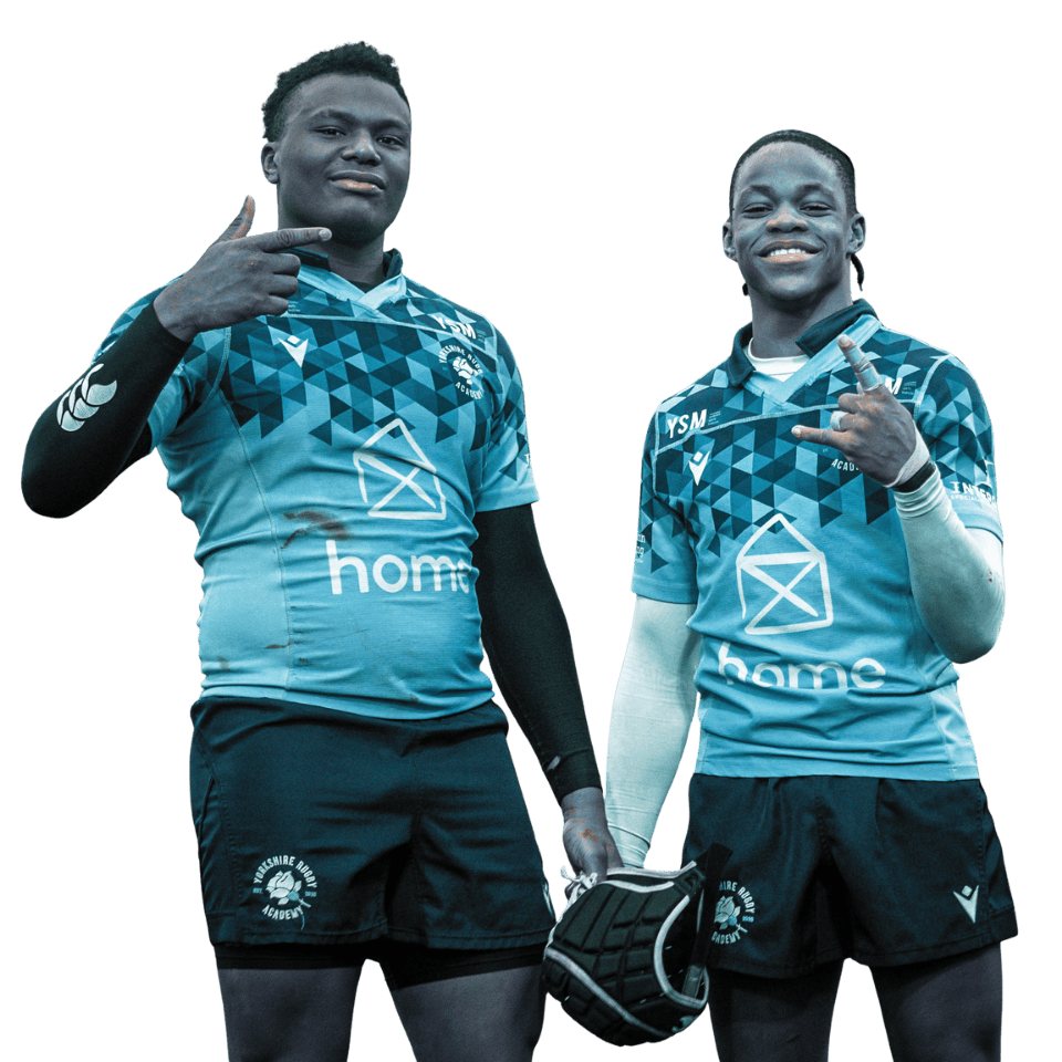 Two rugby players in blue jerseys posing, one pointing at his head and the other making a hand gesture, both smiling, standing against a black background.