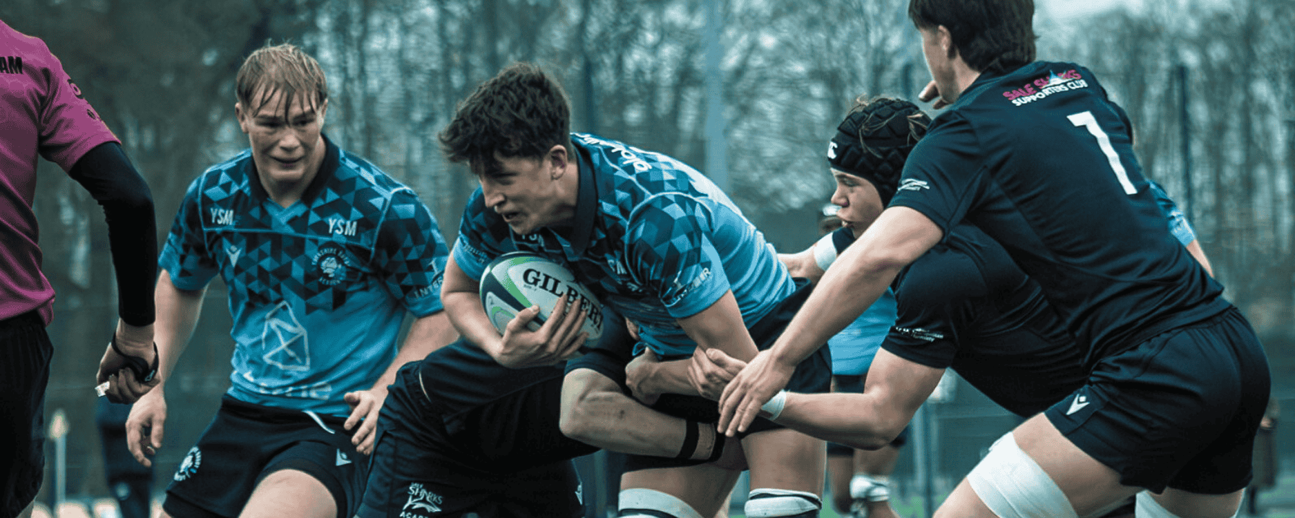 Rugby players in action during a match, with two players in blue jerseys and two players in black jerseys, one of the players in blue holding a rugby ball.