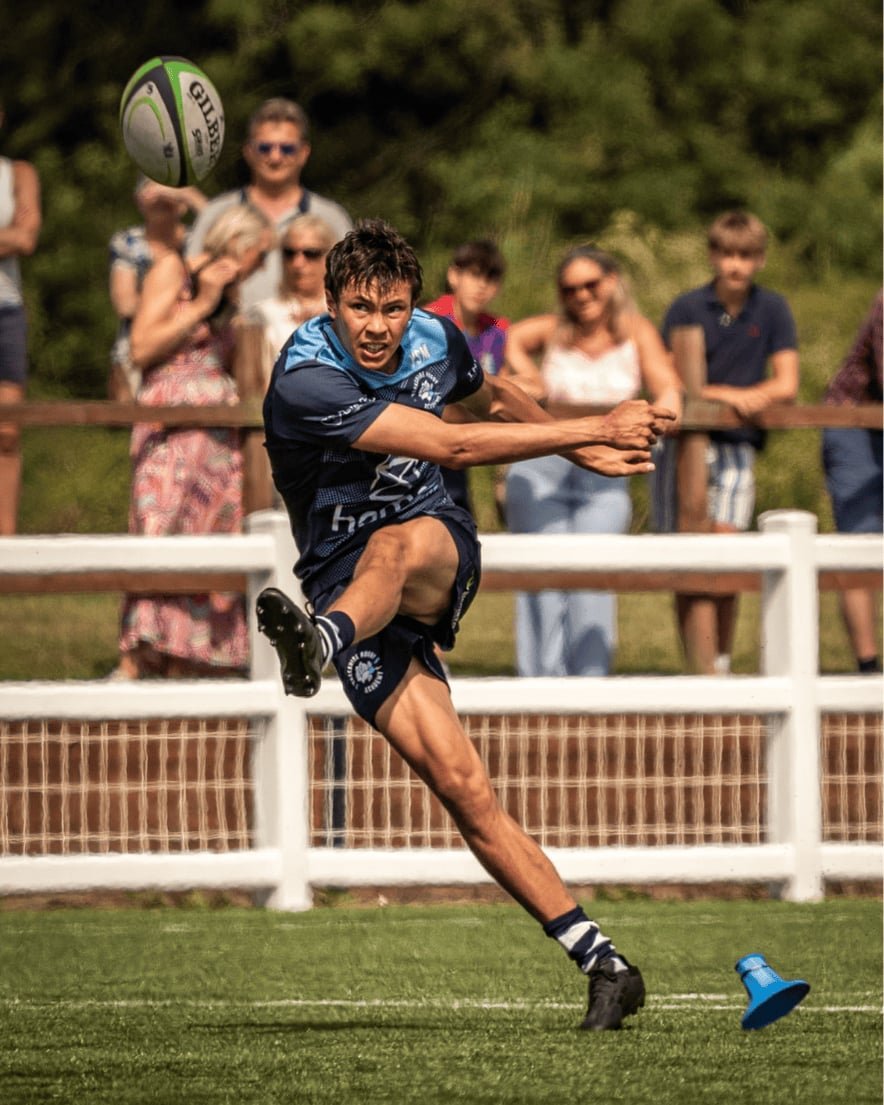 Rugby player kicking a rugby ball on the field while spectators watch from behind a white fence.