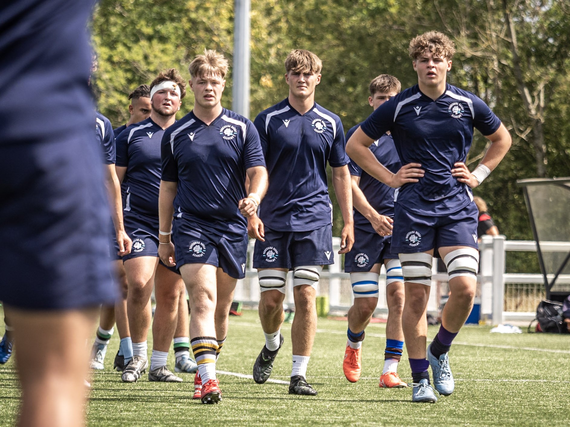 Group of rugby players walking on the field during a game, wearing navy blue uniforms with white accents and various colored socks, with trees in the background.
