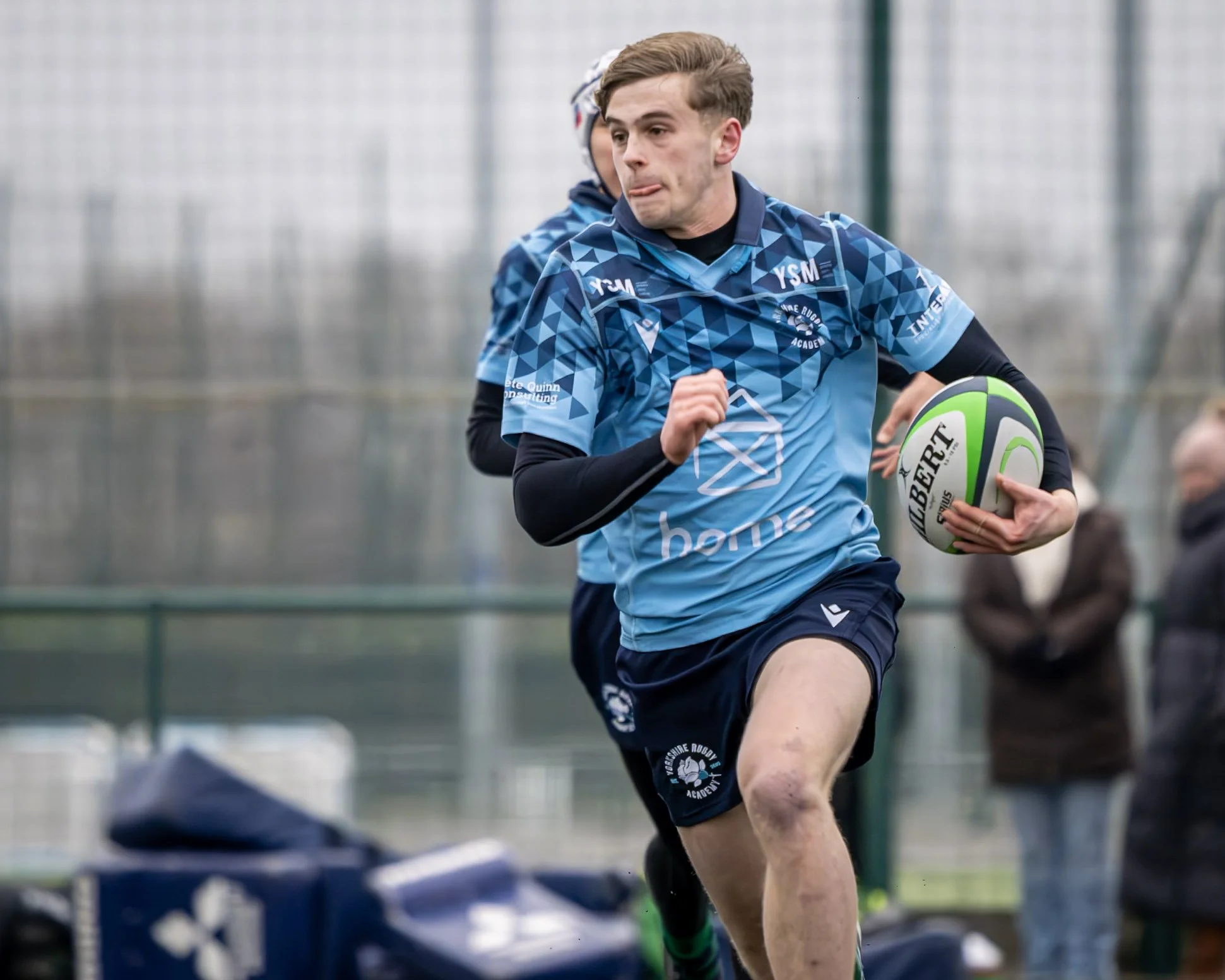 A young man in a blue rugby uniform running while holding a green and white rugby ball on a field, with onlookers nearby.