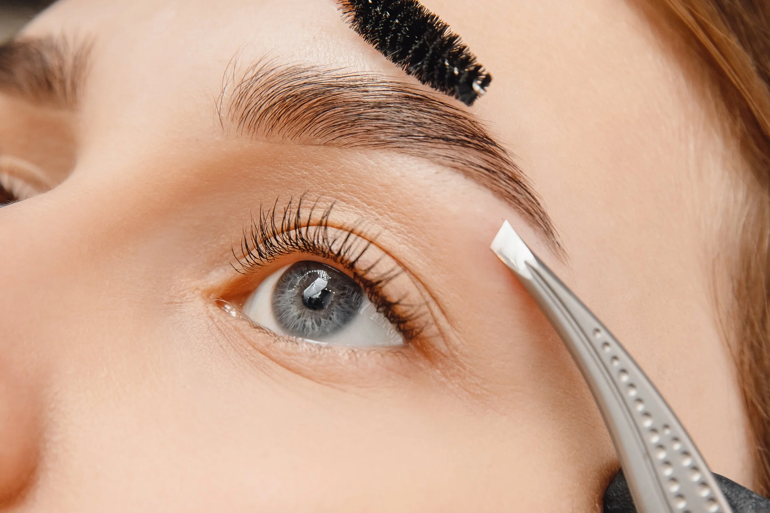 A person with blue eyes has their eyebrow groomed with tweezers by a professional in a cosmetology setting.