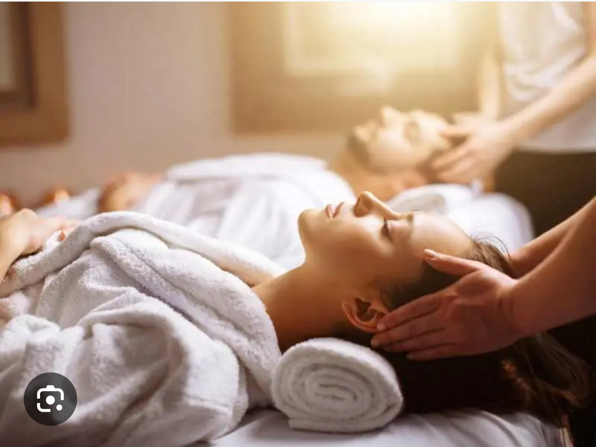 A woman receiving a head massage while lying on a bed with a white towel wrapped around her head in a spa-like setting.