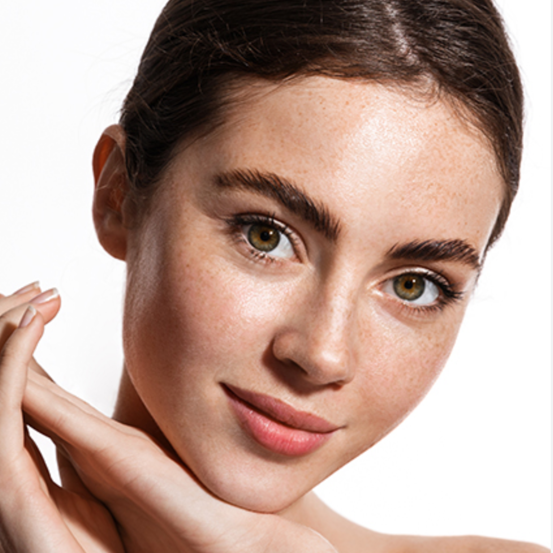 Close-up of a young woman with fair skin and brown hair, smiling softly, with her hand gently touching her face.